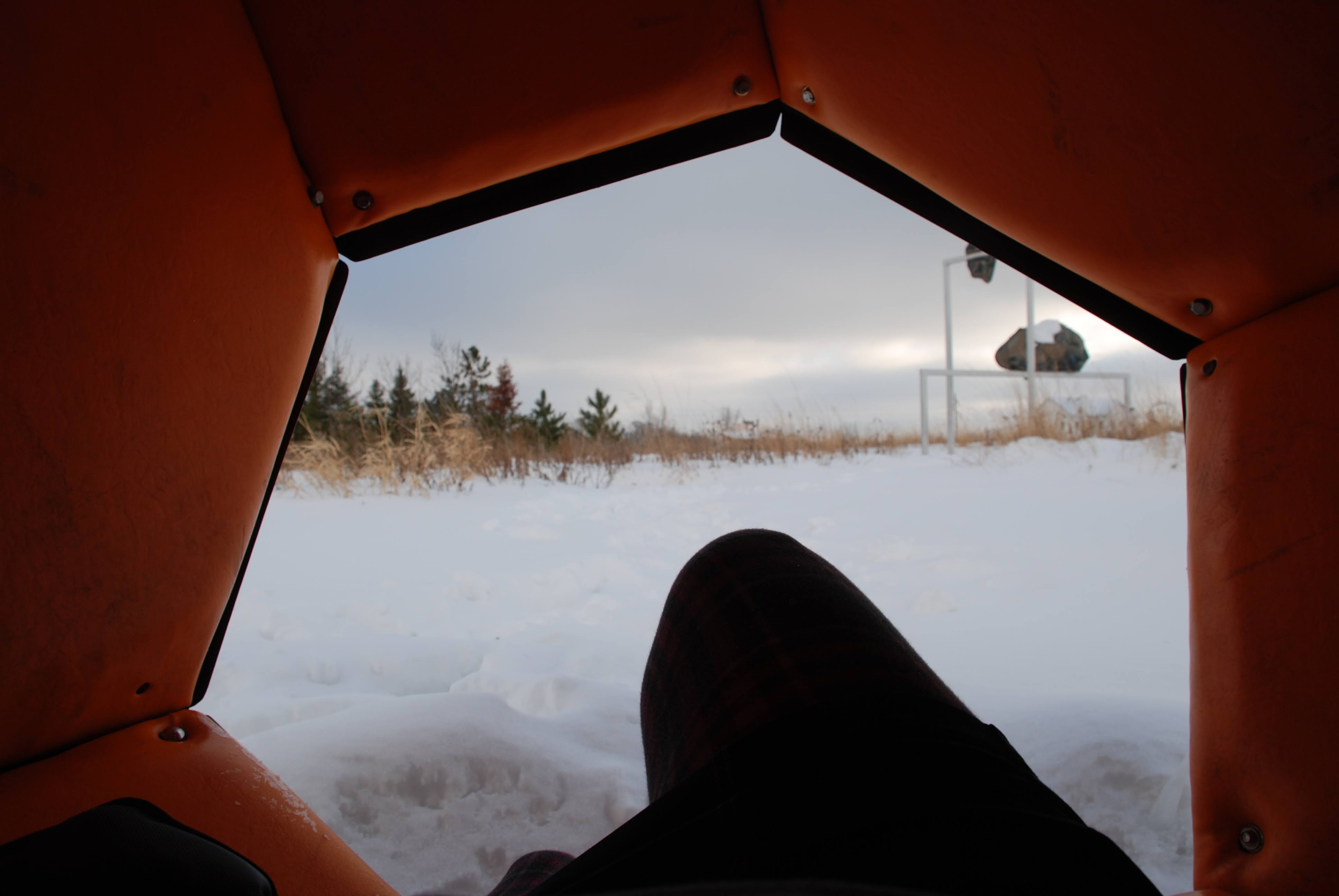 A woman resting and staying warm inside a hexagonal padded sculpture which is orange from the inside. The woman is dressed in all black and the view looks over her raised knee to look out on additional sculptures and the park, covered in snow.